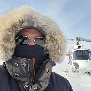 A man takes a selfie in snow clothes in front of a helicopter