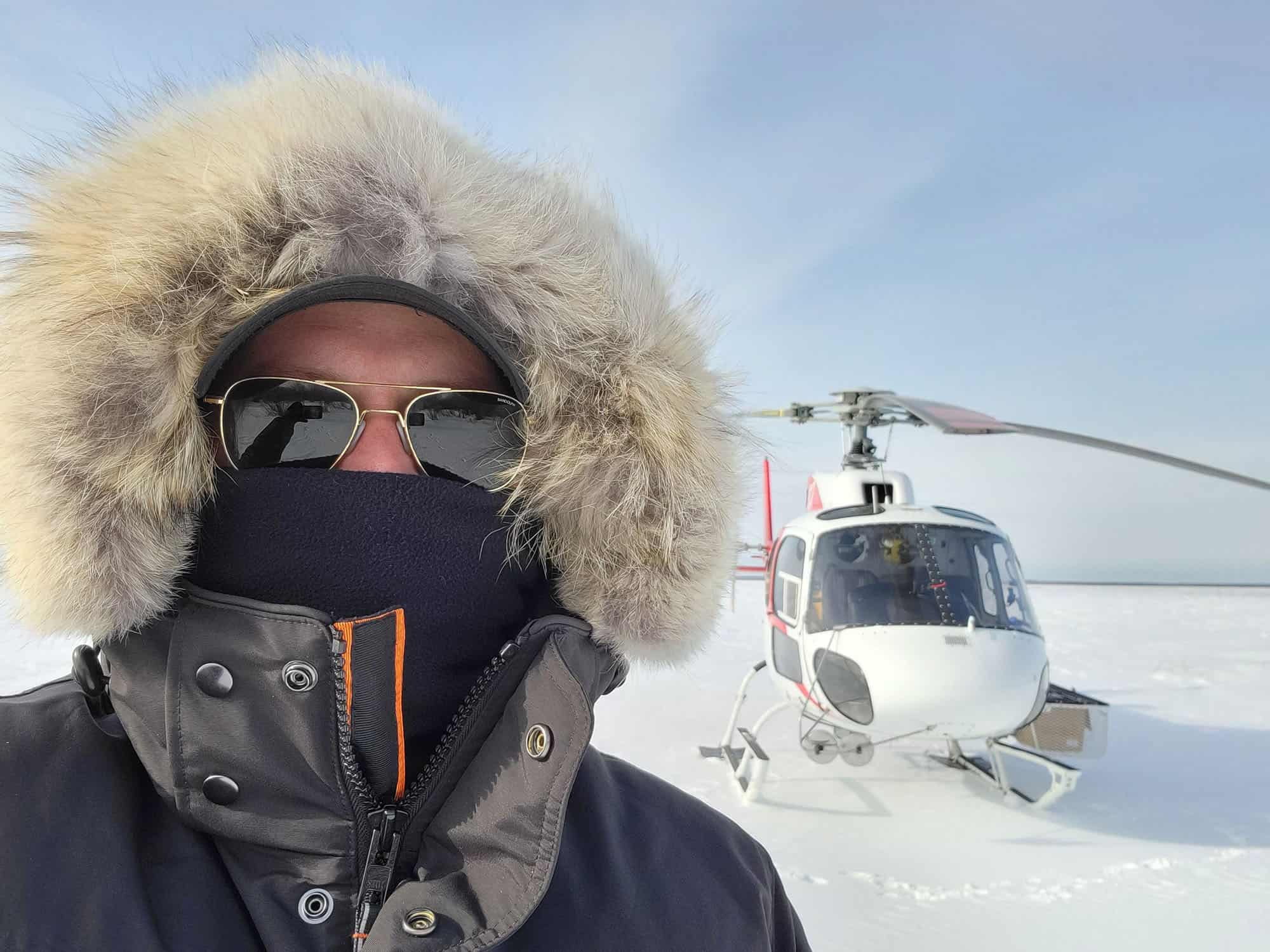 A man takes a selfie in snow clothes in front of a helicopter