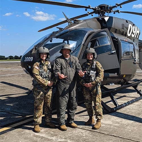 Three smiling people in military fatigues and flight helmets stand in front of a helicopter