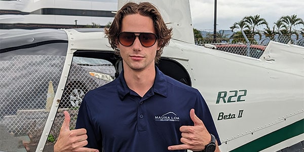 A man stands in front of a helicopter, giving shaka signs with both hands