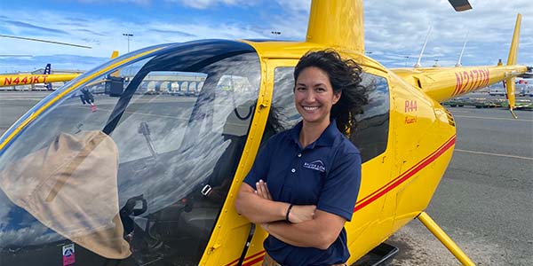 A smiling person stands in front of a helicopter with their arms crossed and their hair blowing in the breeze.