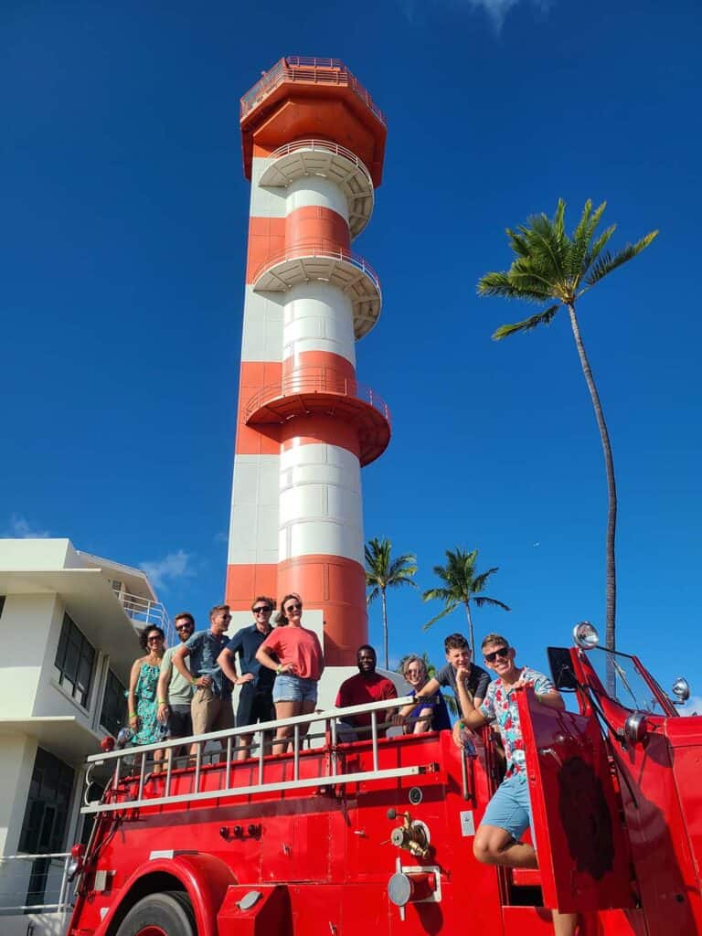 A group of people sit on a old styled firetruck in front of of a large red and white tower.