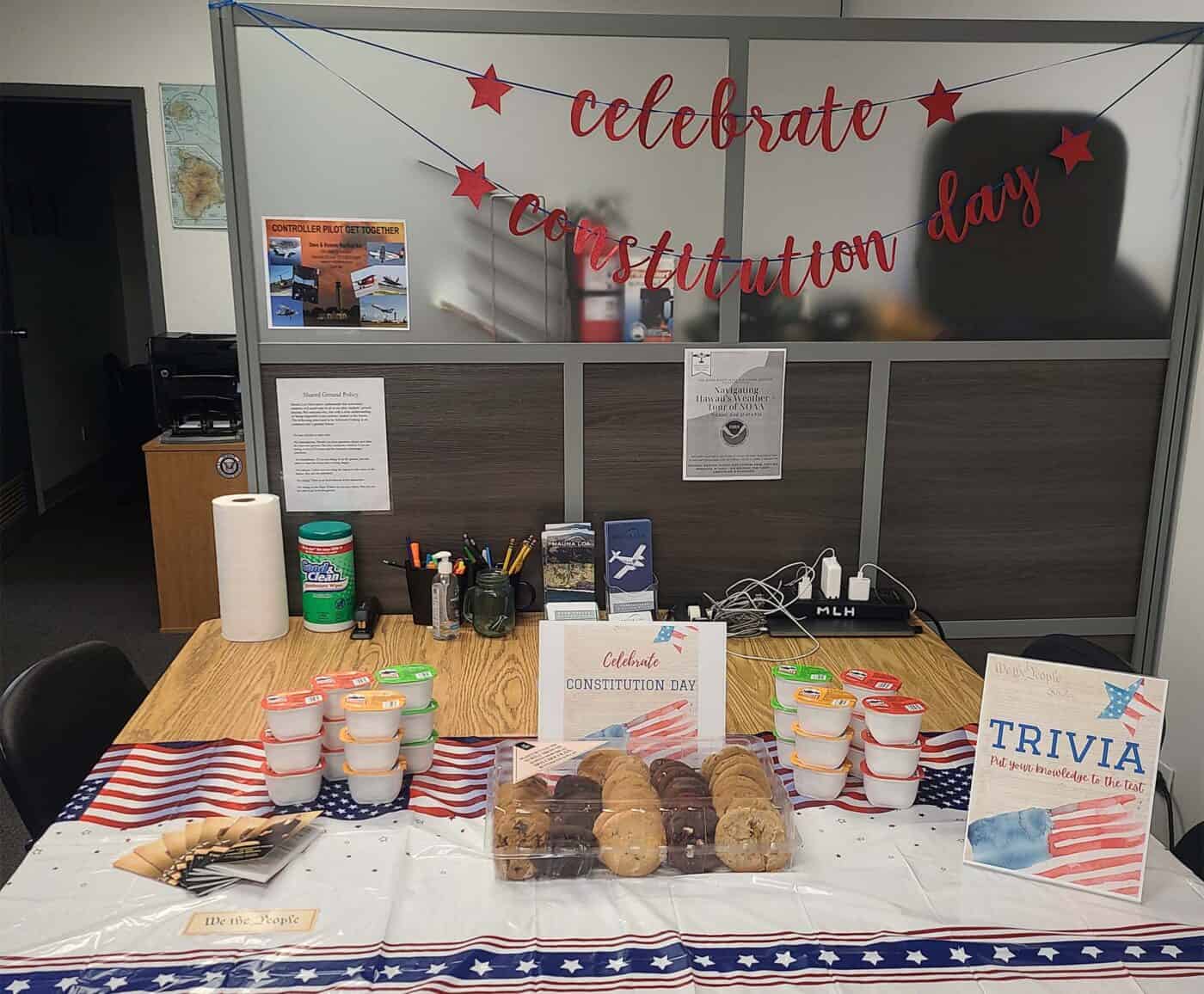 Several snacks sit out on a decorated table. There's a flyer that says "celebrate constitution day", a card that says "we the people", a flyer that says "trivia", and several booklets titled "The Constitution of the United States" artfully displayed. In the background is an office cubicle with a banner saying "celebrate constitution day"