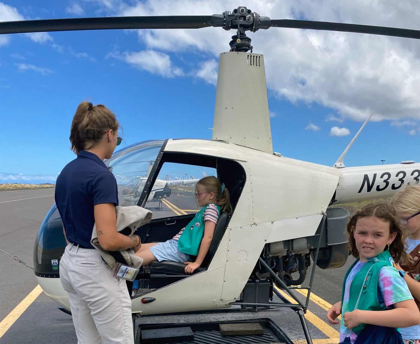 A girl scout sits in the pilot seat of a helicopter while an instructor speaks to them. Several other girl scouts wait in line.