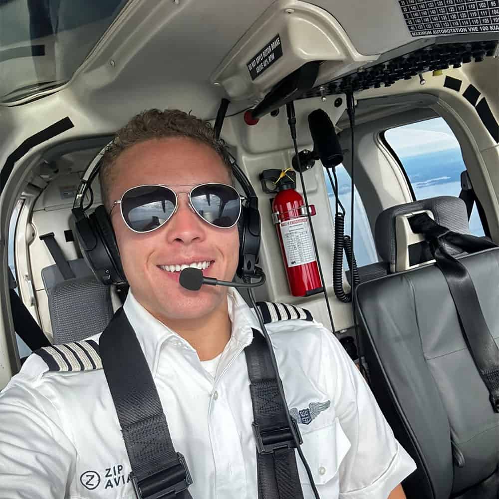 A smiling man wearing sunglasses and pilot's headgear sits in a flying helicopter.