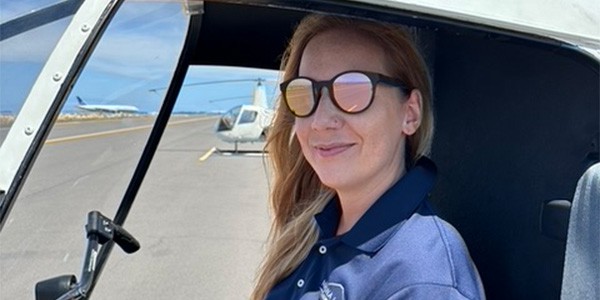 A smiling woman with mirrored sunglasses sits in the pilot seat of a helicopter