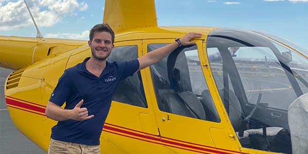 A smiling man leans against a helicopter and gives a shaka sign