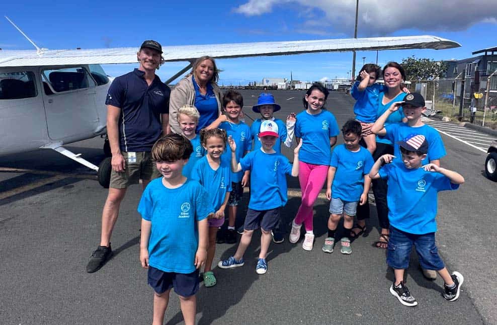 A group of young students and a couple of adults stand in front of a parked airplane.