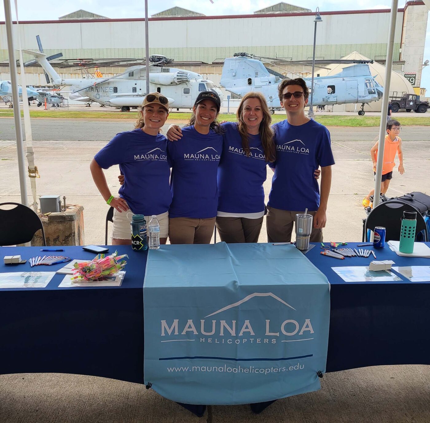 A group of smiling people stand behind a decorated table. A couple of helicopters are parked in the background, and a child plays among them.