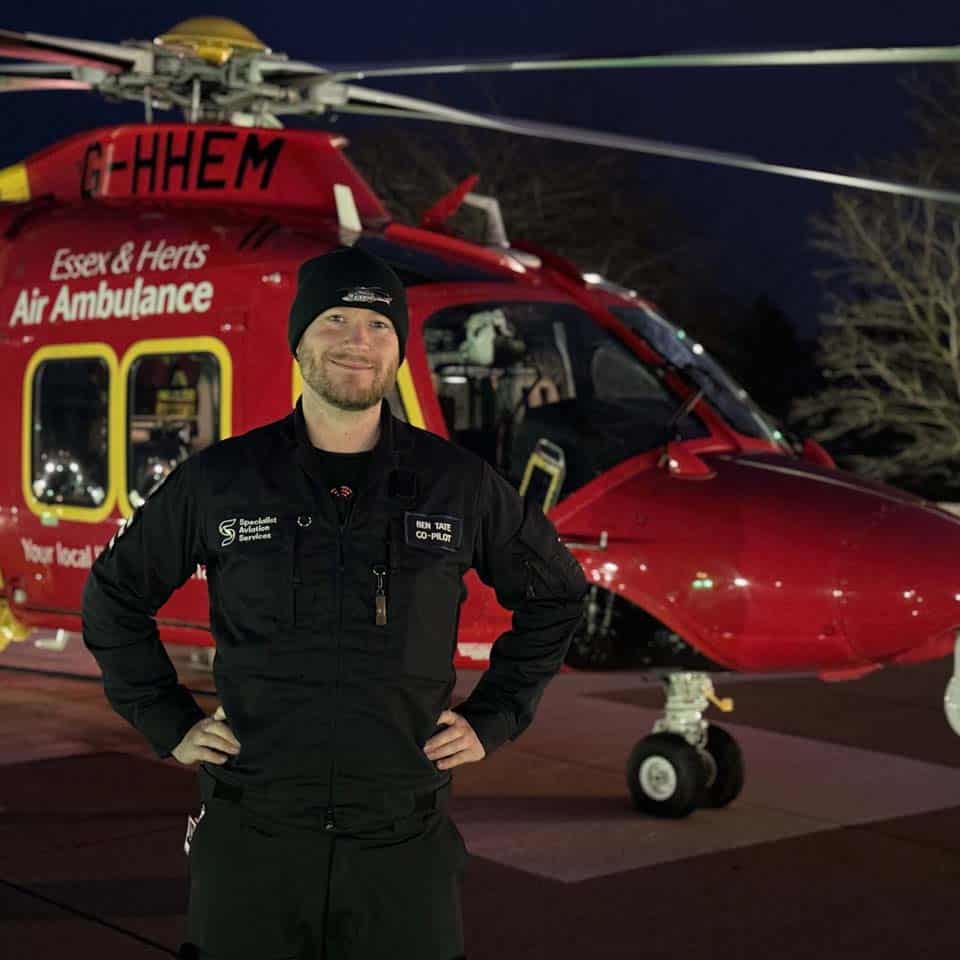 A smiling man in a flight suit stands in front of an air ambulance helicopter with his hands on his hips.