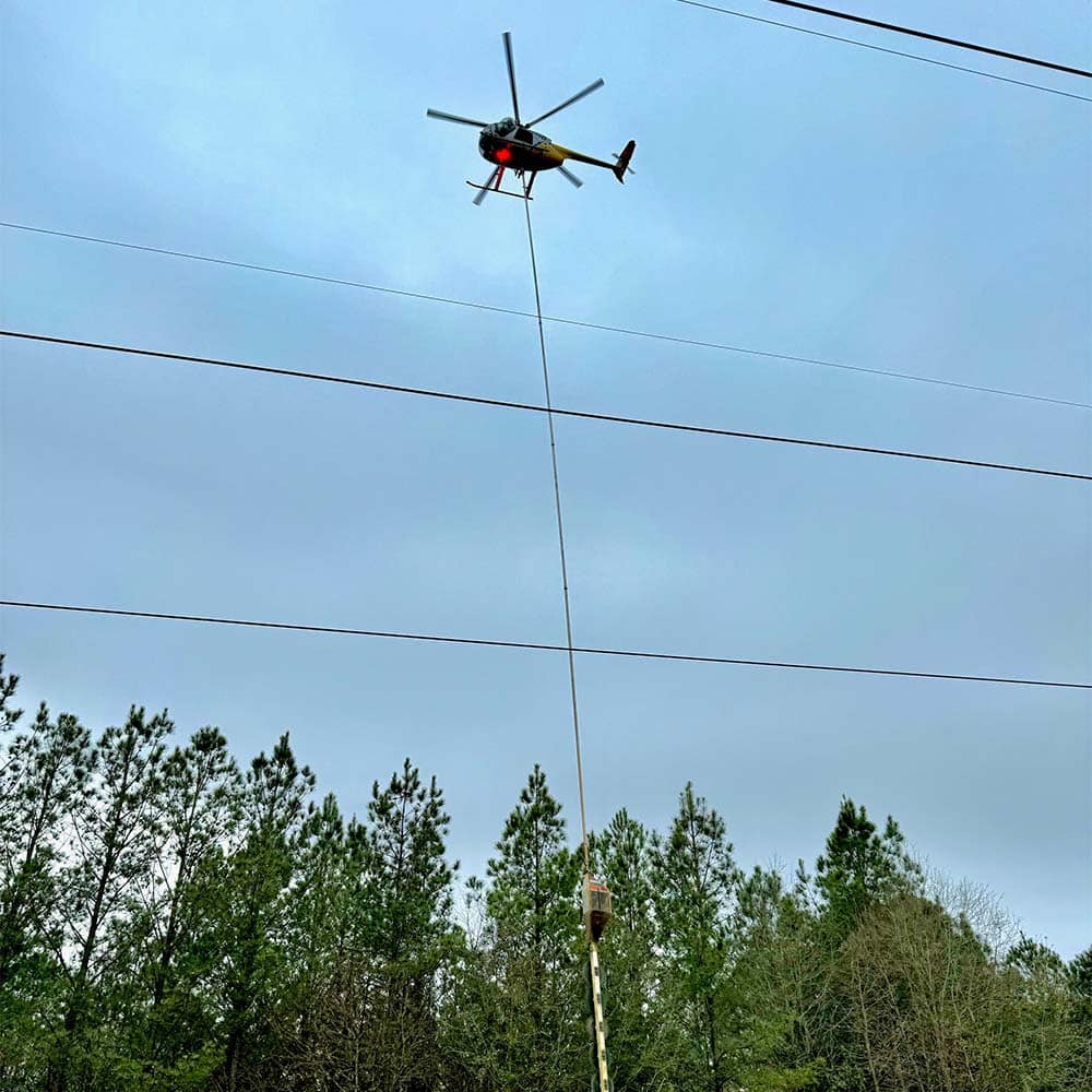 A helicopter flies above some trees. Below the helicopter is a large chainsaw on a line that the helicopter is using to trim the trees.