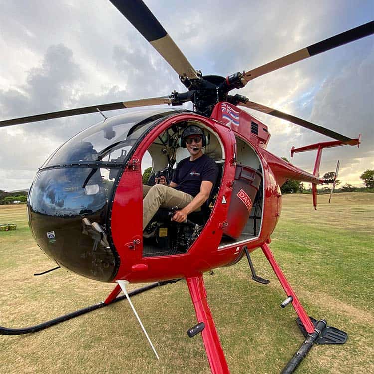 A smiling man sits in the pilot seat of a parked helicopter