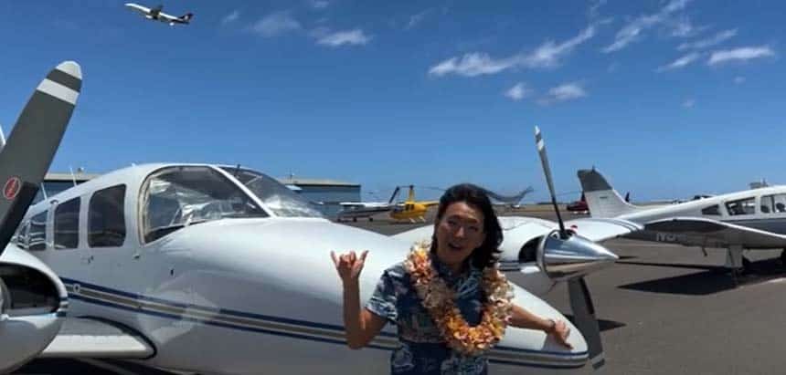 A smiling woman with a lei stands in front of an airplane and gives a shaka sign. A large airplane takes off in the background.