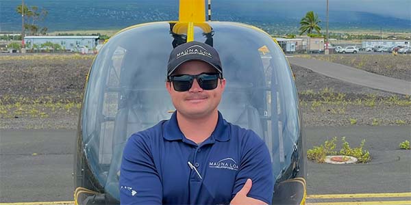 A smiling man wearing sunglasses and a hat stands in front of a helicopter.