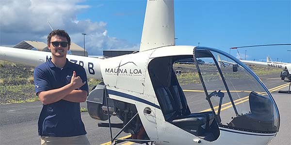 A smiling man stands in front of a parked helicopter and gives a shaka sign