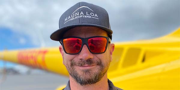 A smiling man wearing sunglasses and a hat stands in front of a helicopter.