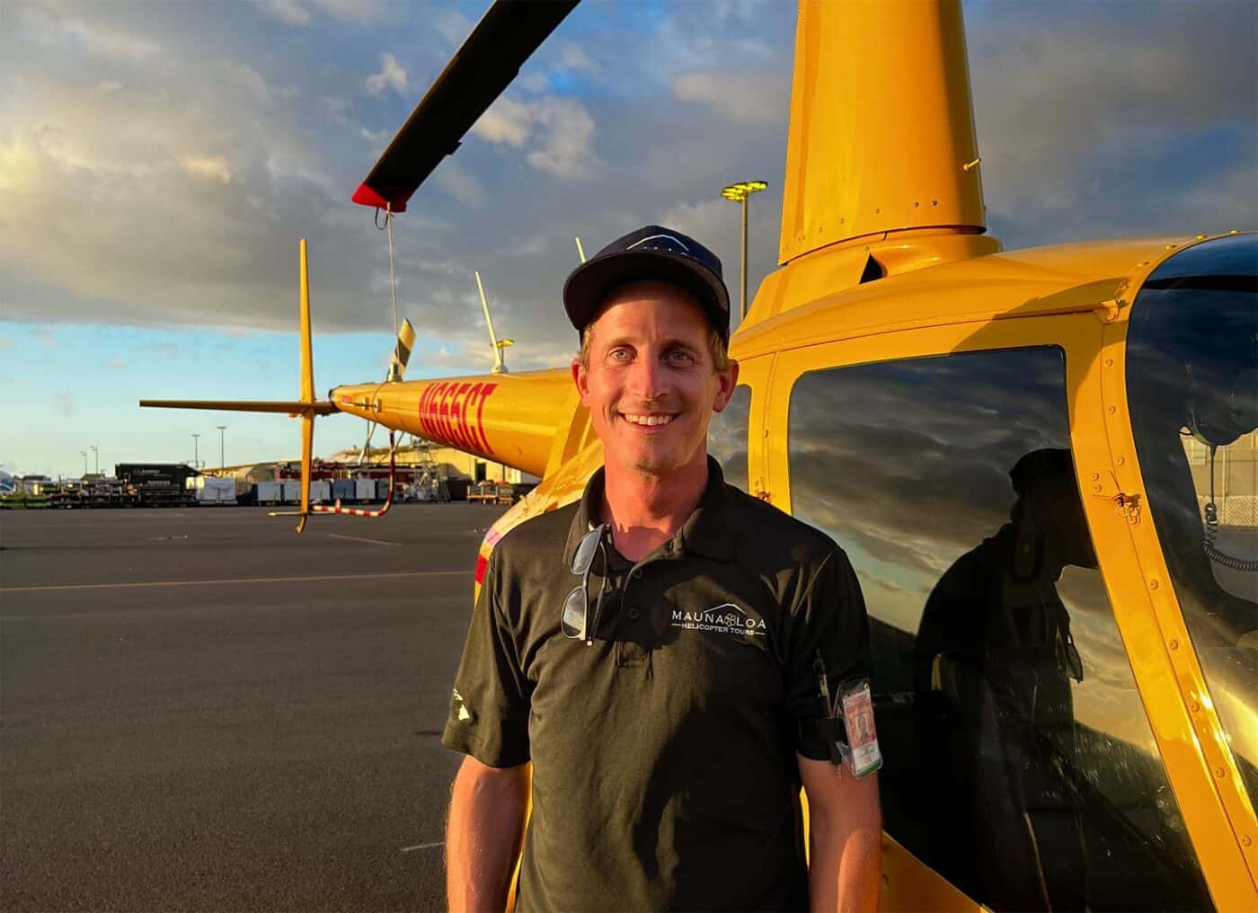 A smiling man stands in front of a helicopter parked on the tarmac. The sun is setting in the background.