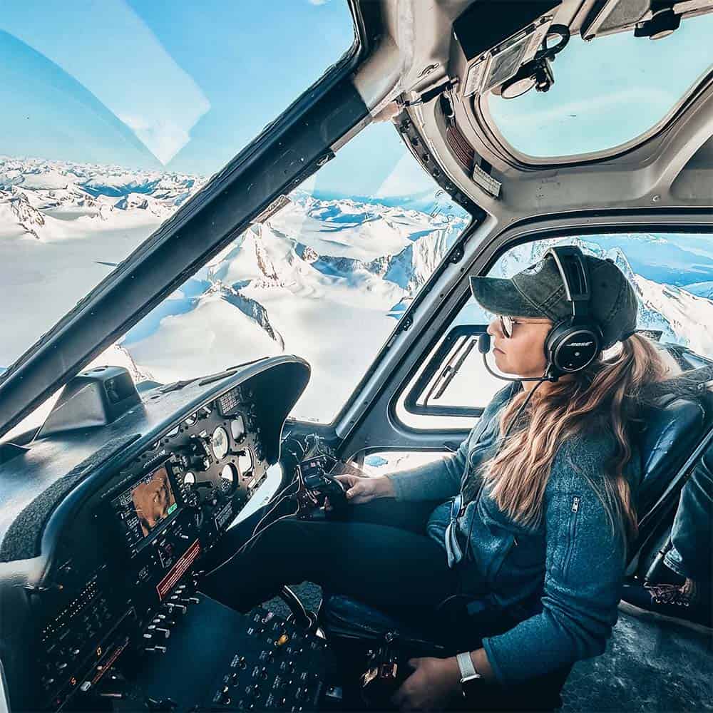 A person pilots a helicopter over picturesque snowy mountains on a clear day