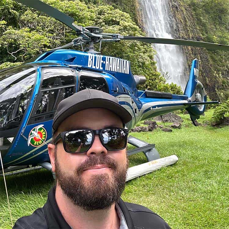 A man takes a selfie in front of a helicopter parked on a hill. A picturesque waterfall flows in the background.