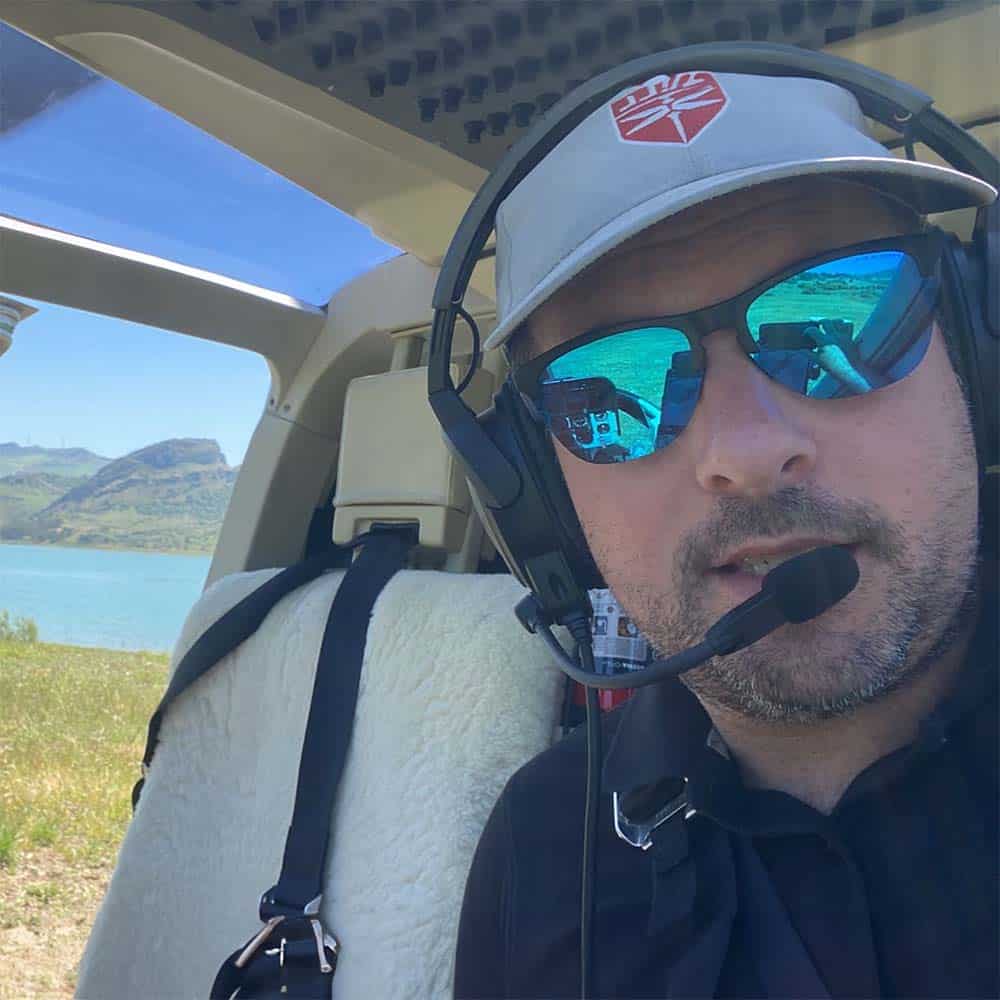 A selfie of a person with sunglasses. They are sitting in the pilot seat of a helicopter, and there is a lake and a mountain in the background.