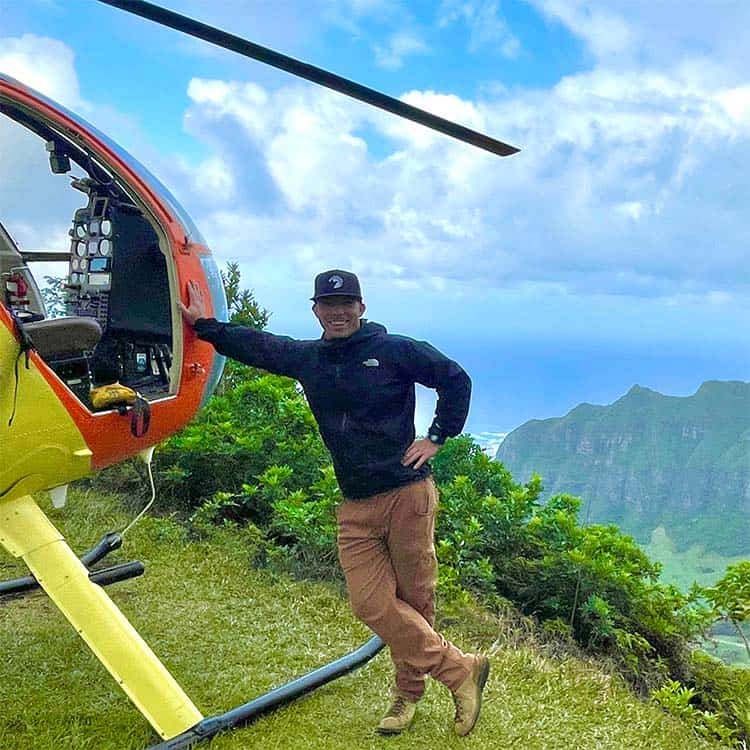 A smiling man leans against a helicopter that is parked on the top of a mountain.