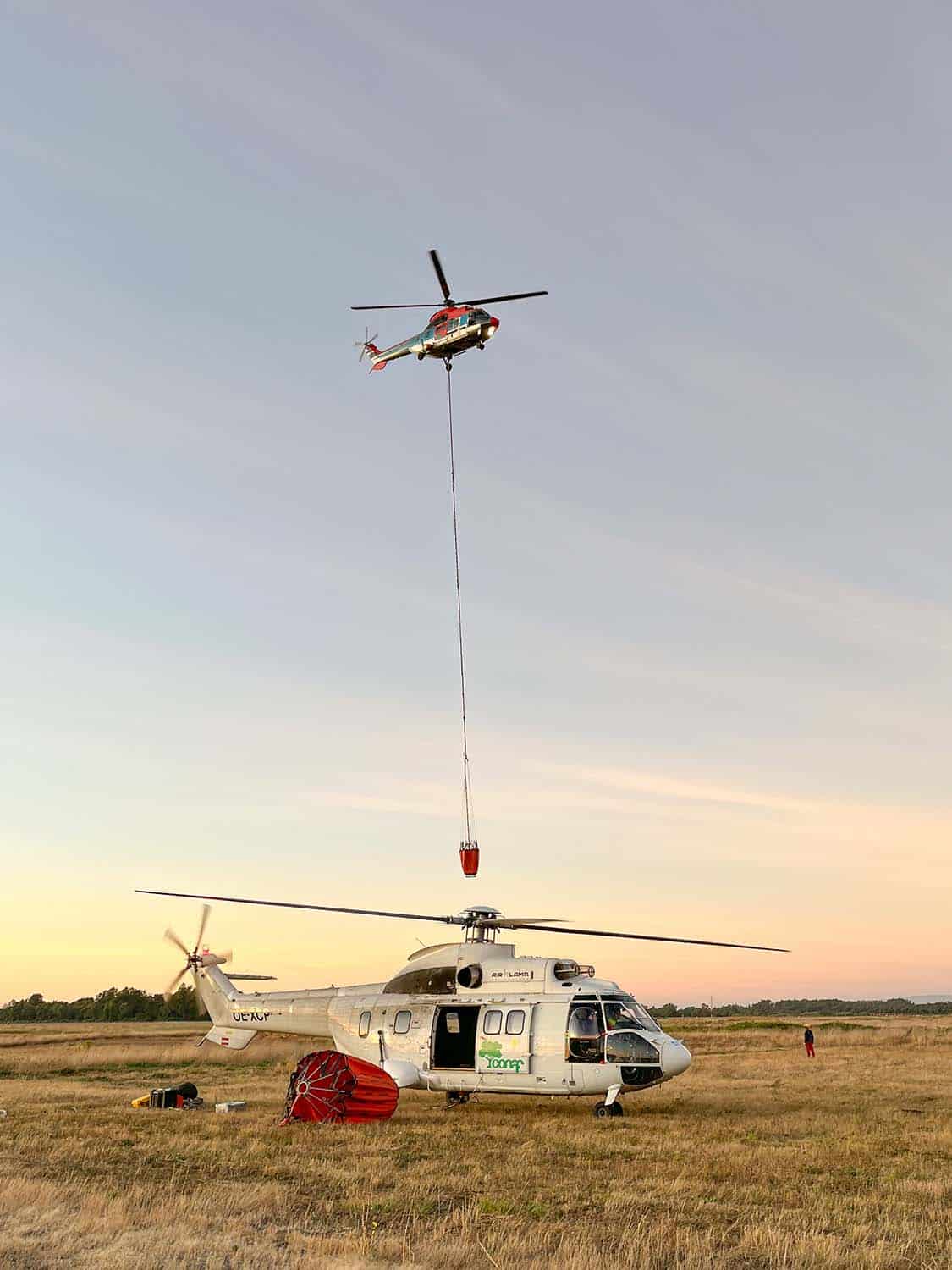 A helicopter flies in carrying a fire bucket at sunset