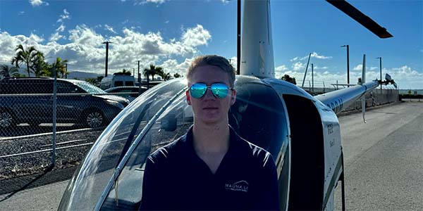 A man with aviator sunglasses stands in front of a parked helicopter on a sunny day