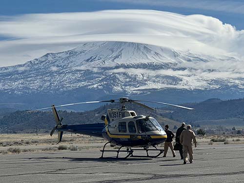 A helicopter sits on a tarmac with a picture-esque snowy mountain in the background. Three people in flight suits walk towards the helicopter