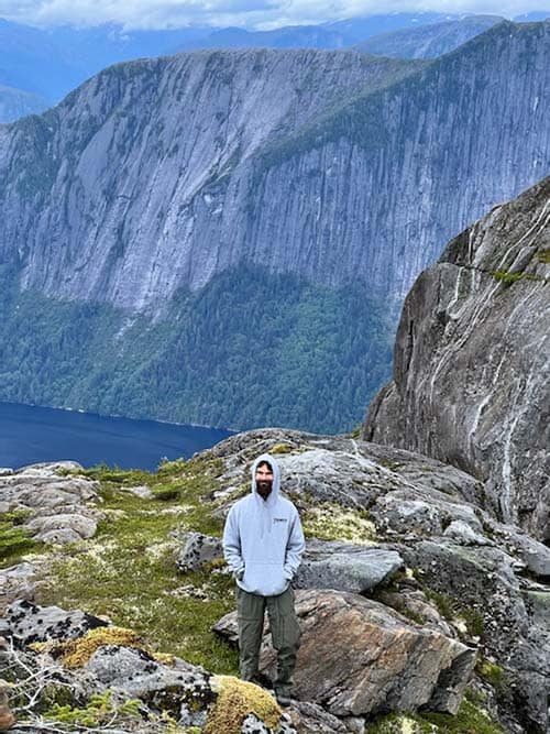 A smiling man in a hoodie stands on a cliff edge. In the background are more cliffs overlooking a lake