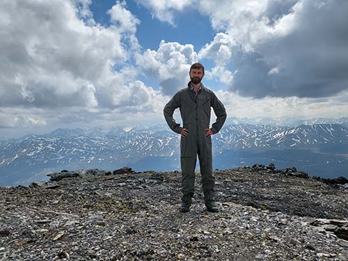 A smiling man in a flight suit stands on a mountain top. in the background are more snowy mountains.