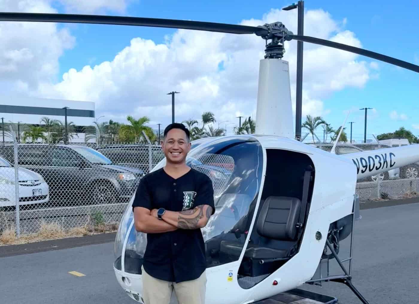 A smiling person stands in front of a parked helicopter with their arms crossed.