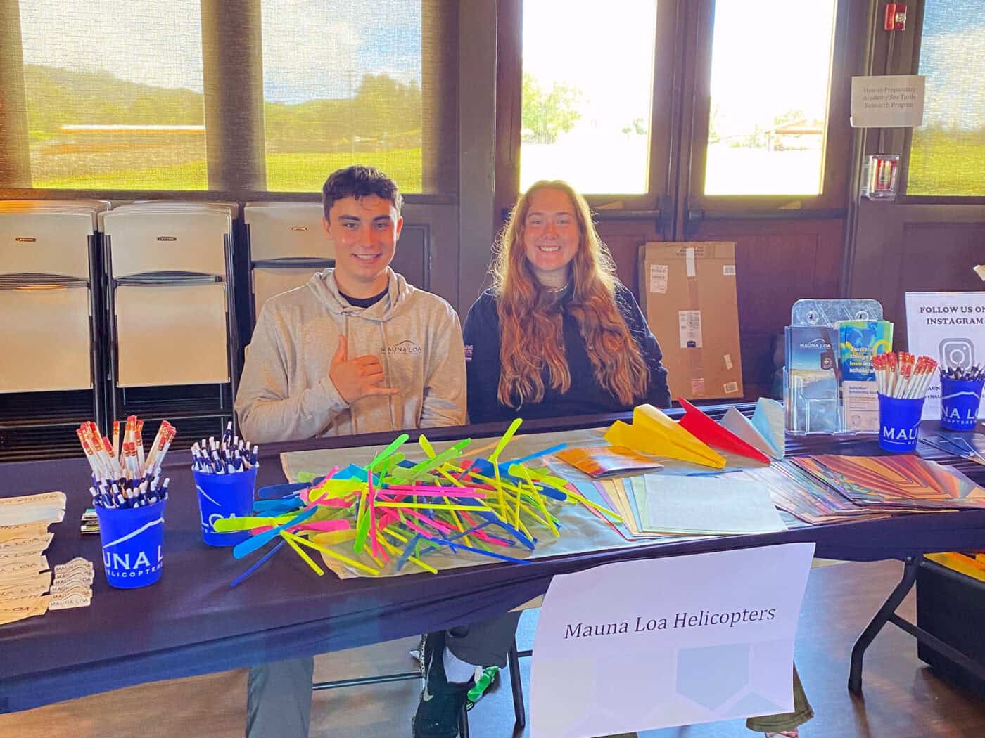 Two smiling people sit at a table with some merch on it. A sign labelled Mauna Loa Helicopters sits on the front of the table. One of the people gives a shaka sign.