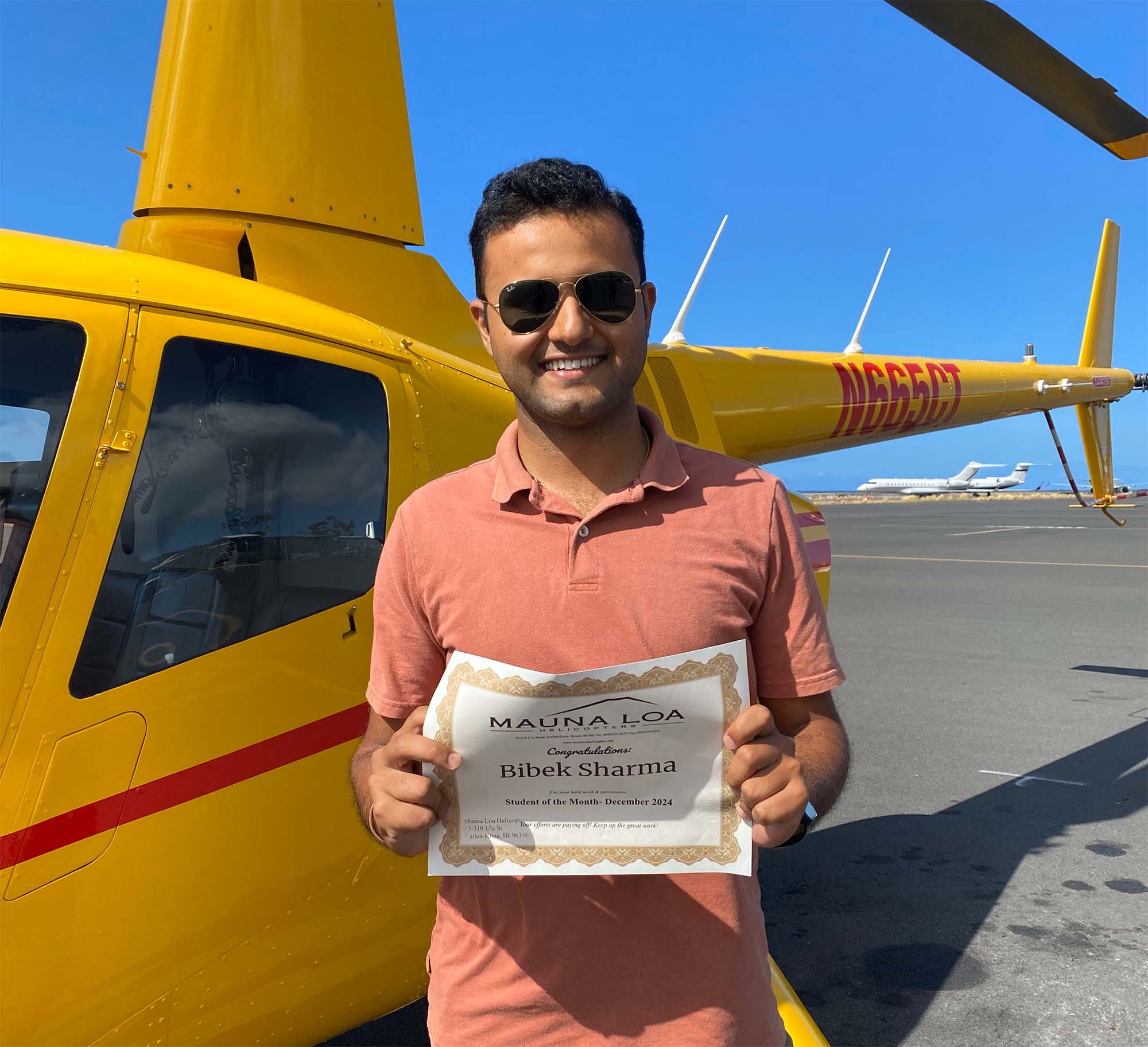 A smiling person stands in front of a helicopter and holds a certificate